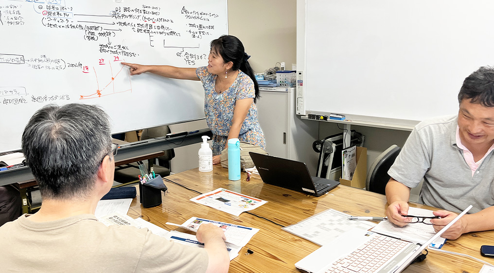 プロジェクトサポートの風景。ホワイトボードに書き込まれた事業スキームやグラフを指し示しながら、専門スタッフが相談者と共に具体的な活動フェーズや戦略を練り上げている様子