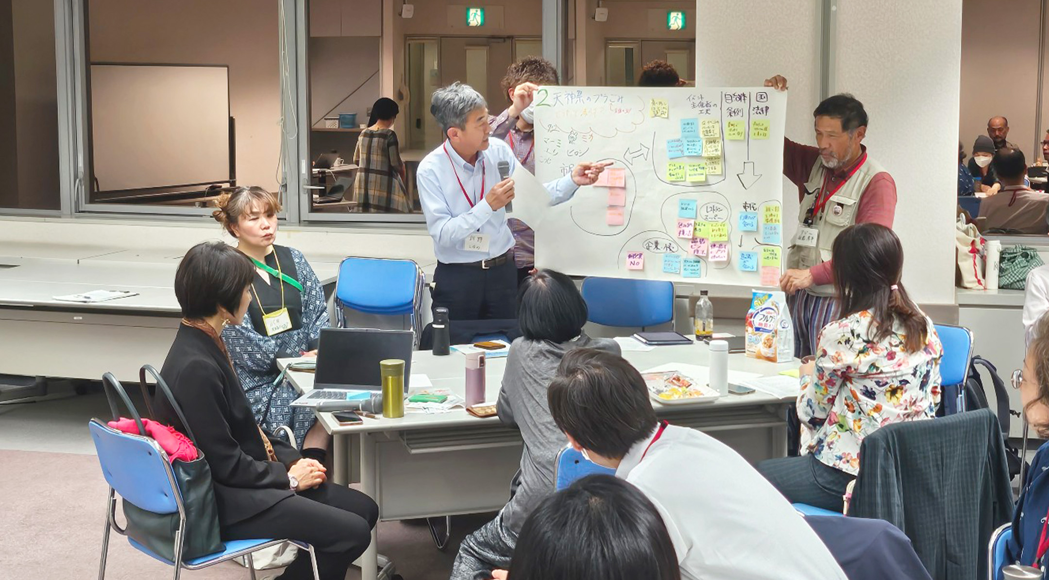 地域課題解決に向けた対話の場の実践。複数の参加者がテーブルを囲み、模造紙と付箋を使って可視化されたアイデアを指し示しながら発表・議論を行っている、双方向の学びのシーン